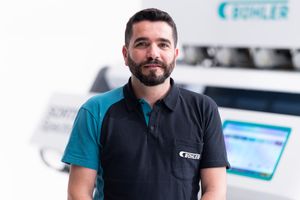 A smiling man with a beard wearing a teal and black polo shirt stands in front of industrial Buhler machinery.