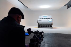 Man in black cap films a large industrial grain sorting machine displayed in a white gallery space.