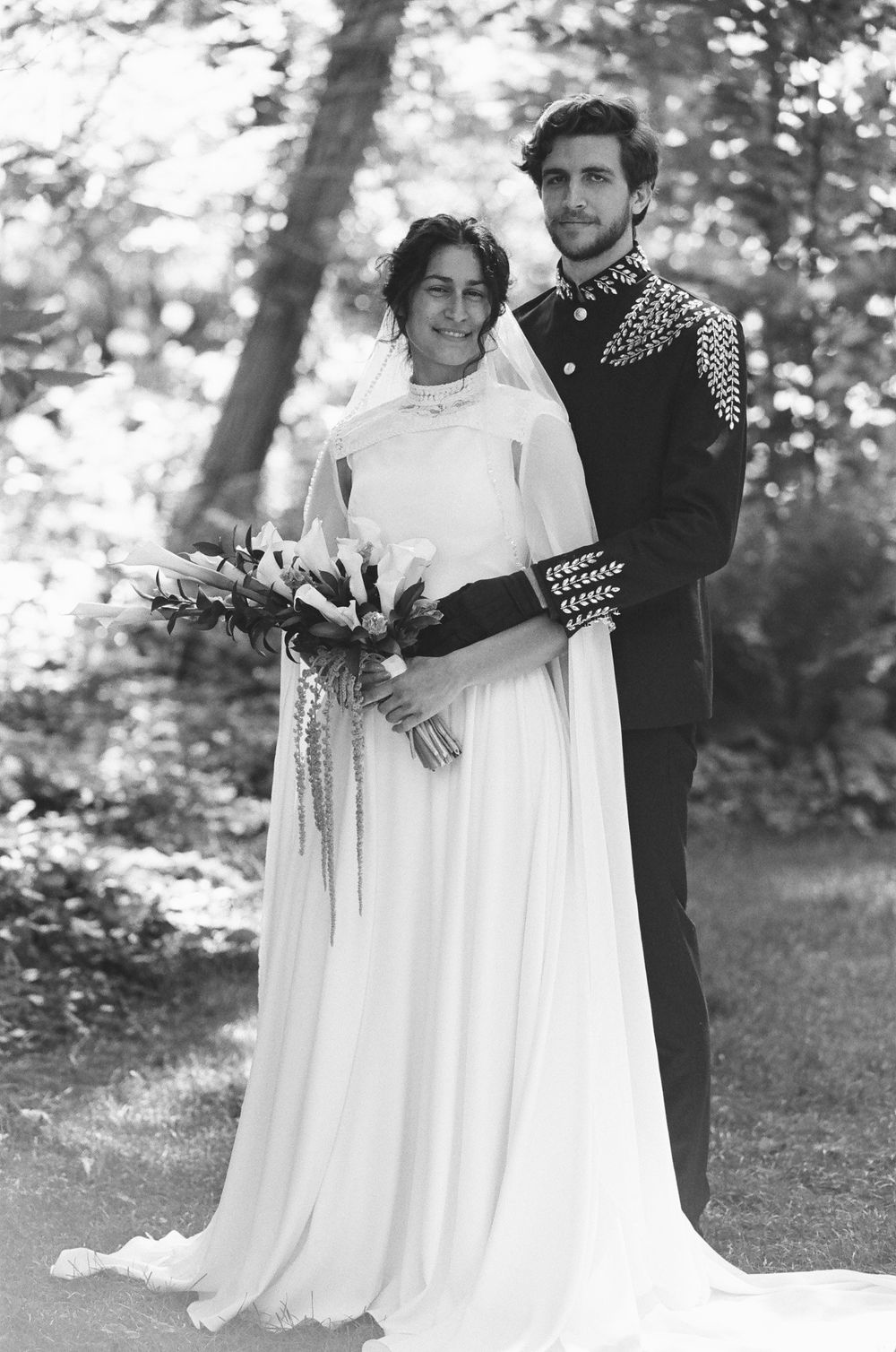 Black and white wedding photo of a bride in a flowing gown with bouquet and groom in military-style jacket outdoors.