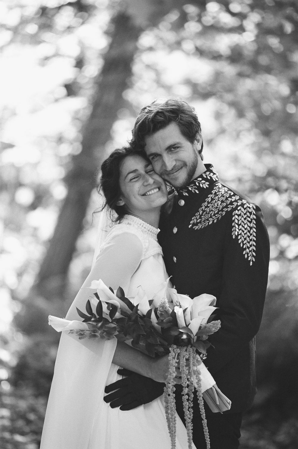 Black and white portrait of smiling bride and groom embracing in a sunlit forest setting.