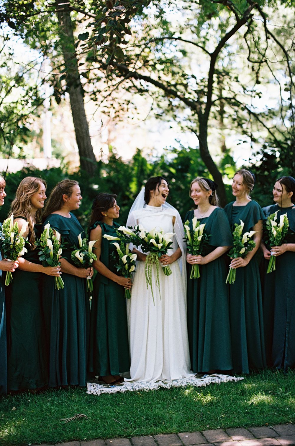 Bride in white gown with veil surrounded by bridesmaids in emerald green dresses holding white calla lilies.