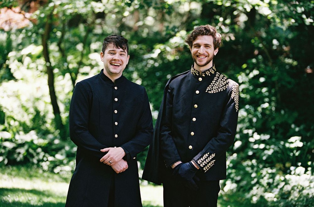 Two grooms in matching black military-style suits with gold buttons smiling outdoors at a wedding.