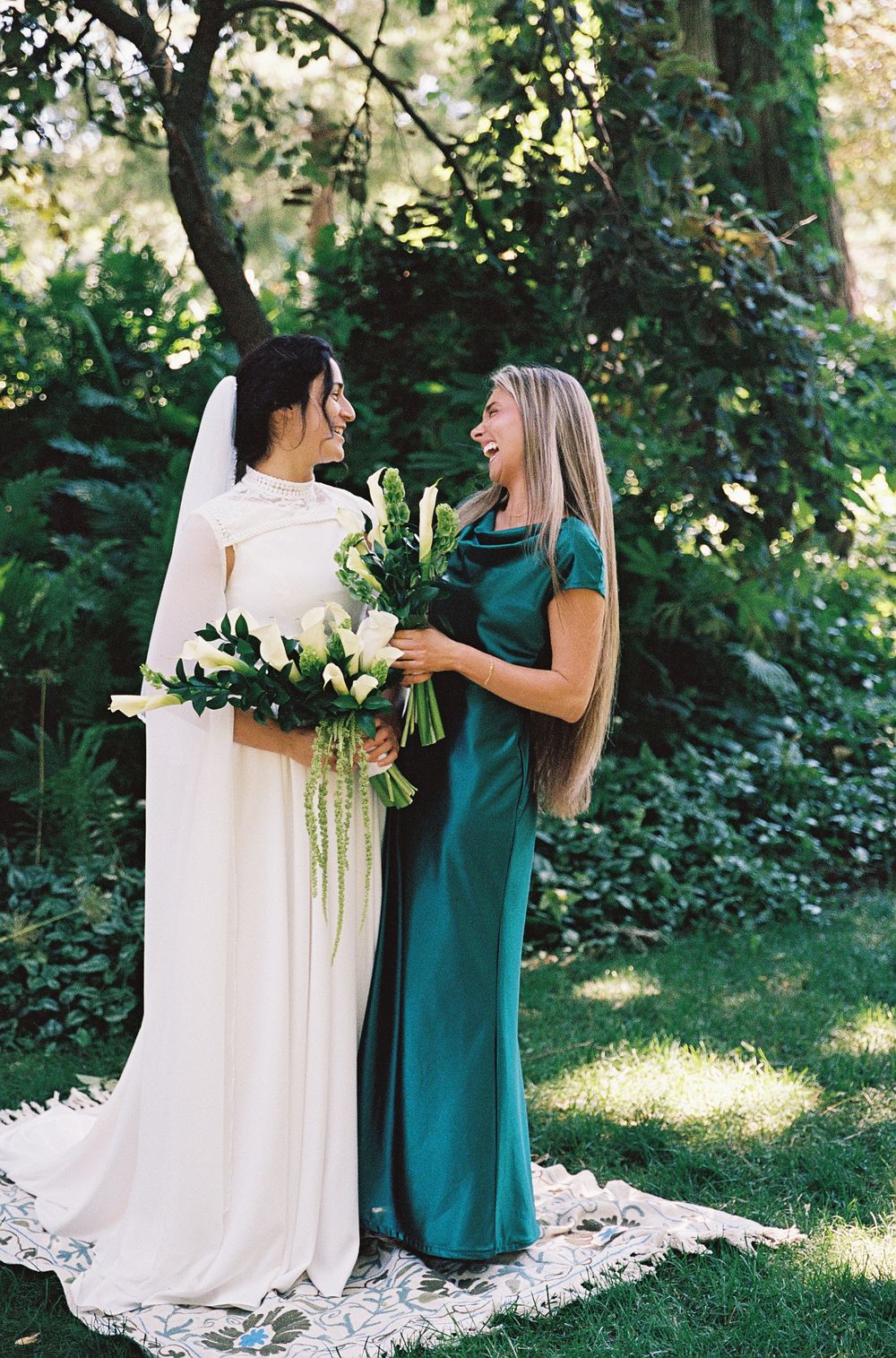 Bride in white gown and bridesmaid in emerald green dress laughing together in garden setting.