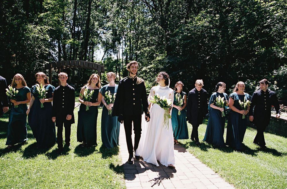Bride and groom stand with full wedding party in emerald and black attire on a sunny garden pathway.
