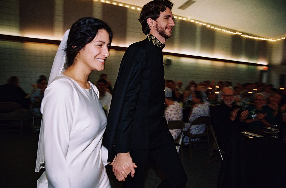 Smiling bride and groom in black suit walk hand in hand at wedding reception.