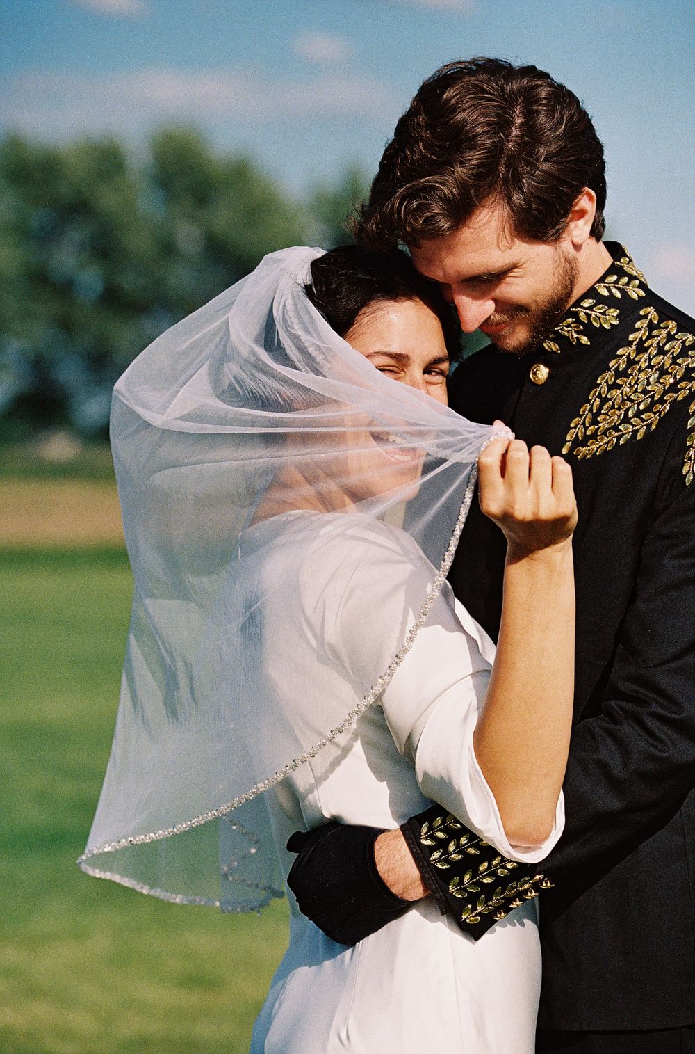 Bride and groom embracing outdoors, her veil lifted, wearing a military-style embroidered jacket at their wedding.