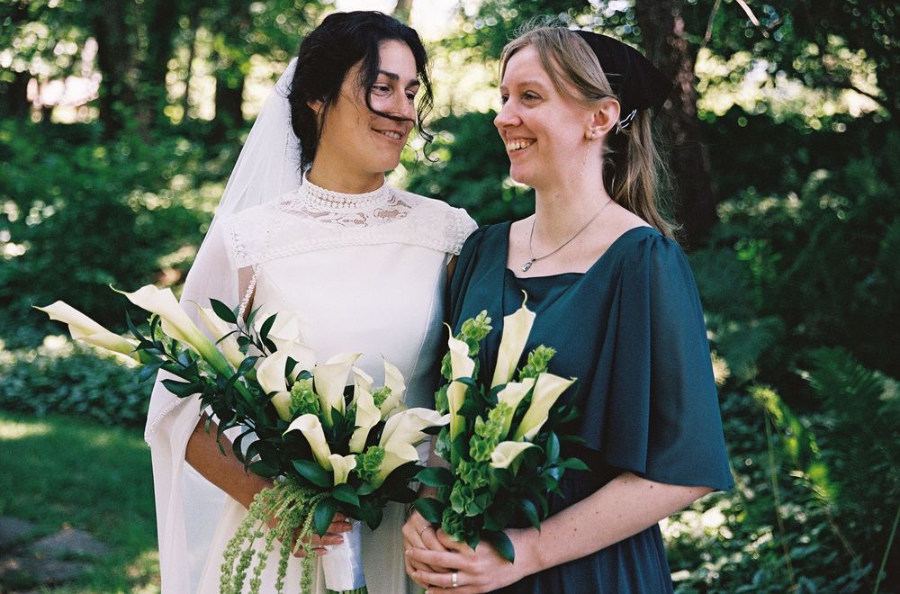 Bride in white gown and bridesmaid in teal dress smile at each other holding calla lily bouquets.