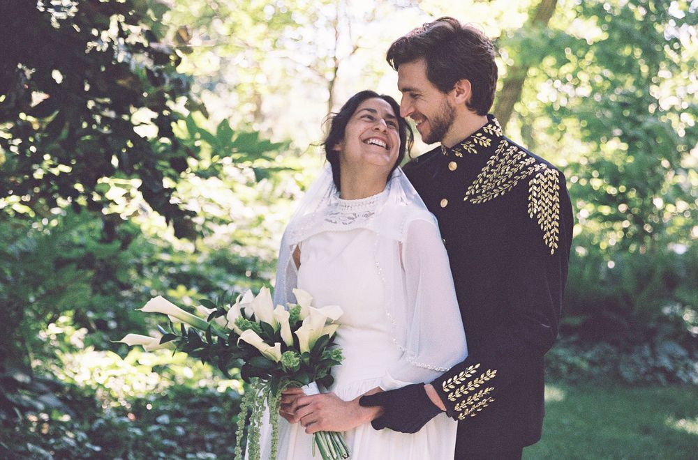 Smiling bride holding white calla lily bouquet embraced by groom in ornate black jacket outdoors.