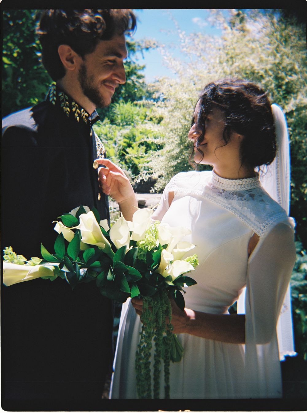 Bride and groom exchange smiles outdoors, she in white high-neck gown holding calla lily bouquet.