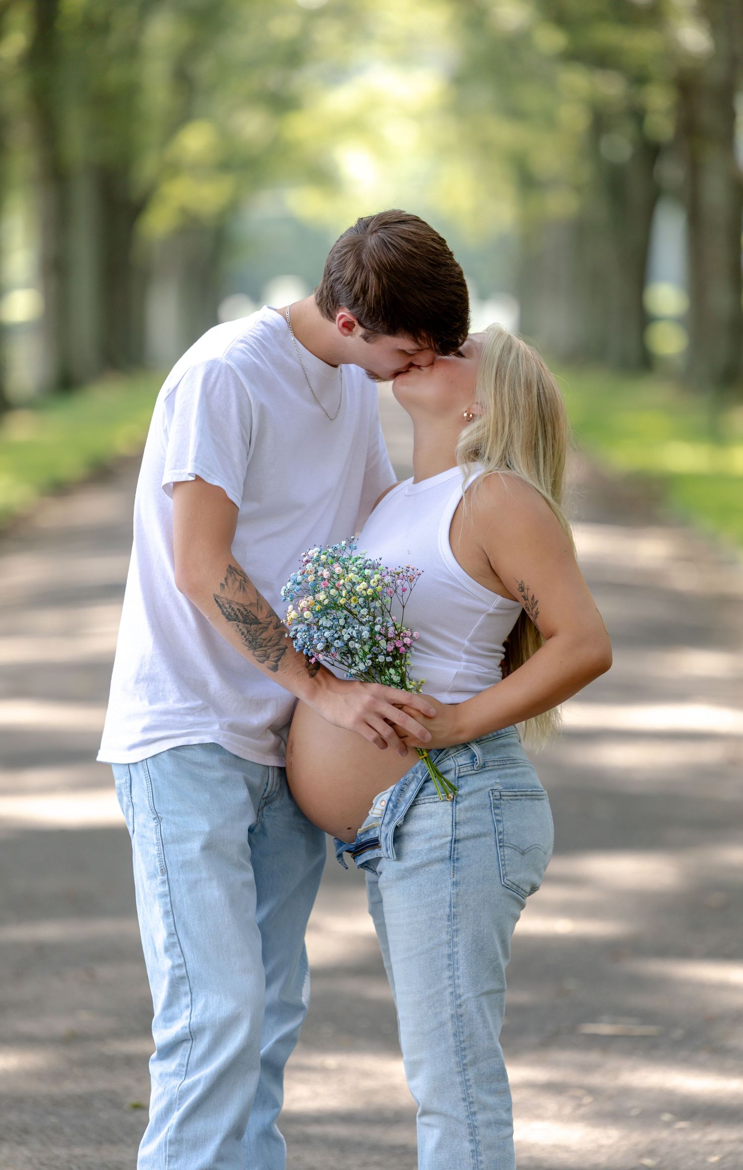 Maternity photographer Rome, GA Couple sharing a kiss on a tree-lined path, woman holding wildflowers with a visible baby bump