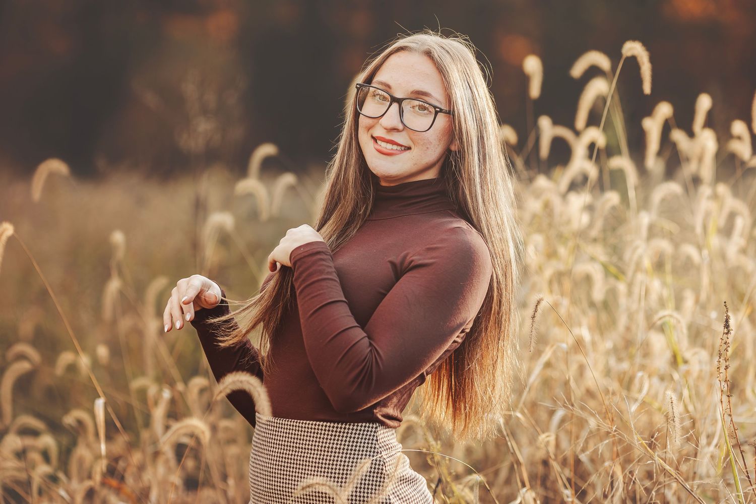 Smiling young woman with long hair and glasses posing in a golden autumn field with tall grass.