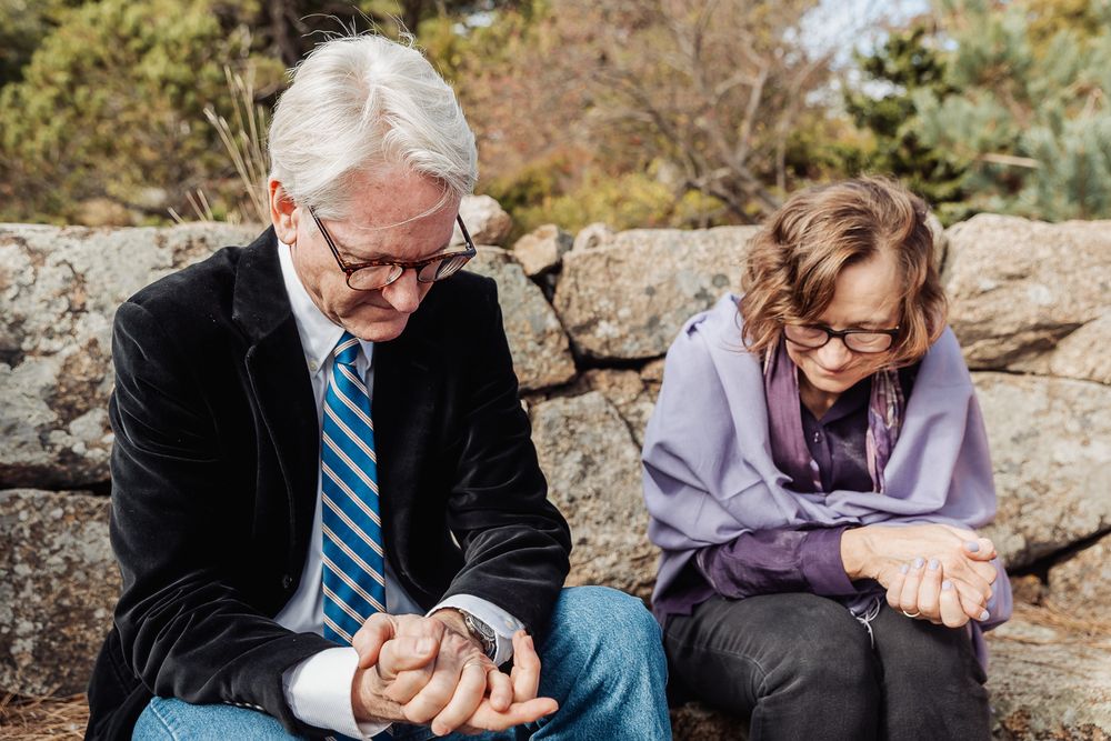 An elderly man and woman sit on stone wall, looking at small object in his hands, outdoors among trees.