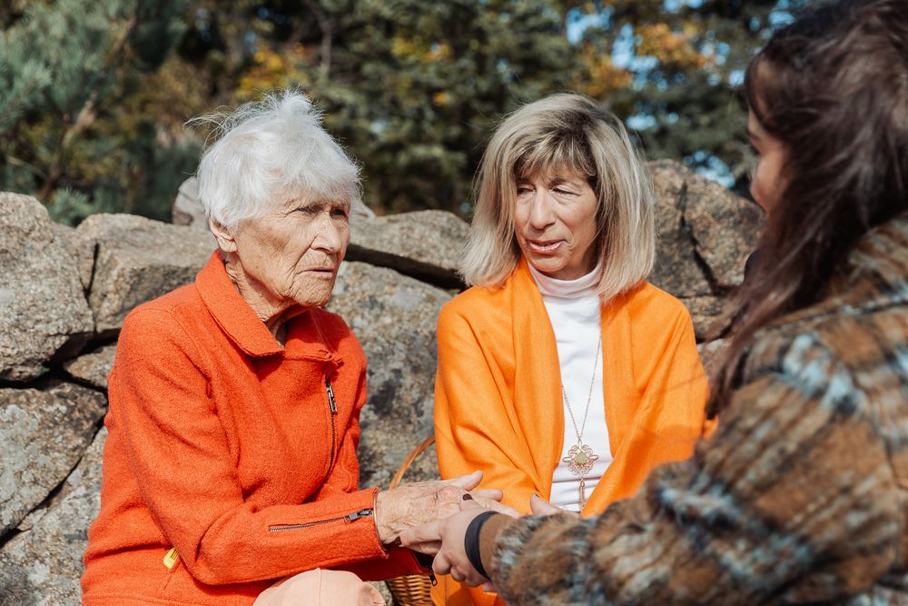 Two elderly women in orange sit on rocks outdoors, holding hands with a younger woman in a warm autumn setting.