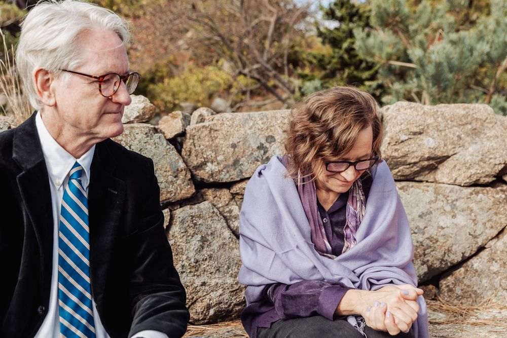Elderly couple sitting on stone wall outdoors, man in suit with blue tie, woman in purple shawl and glasses.