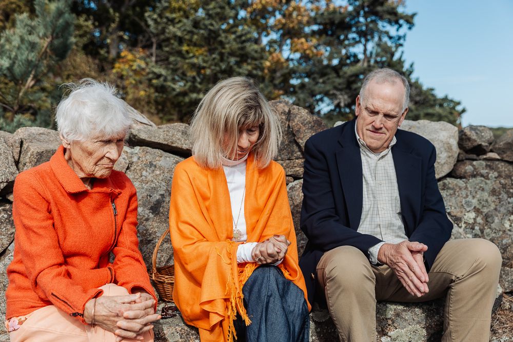 Three elderly people sitting on a stone wall outdoors, wearing orange and navy clothing on a sunny day.