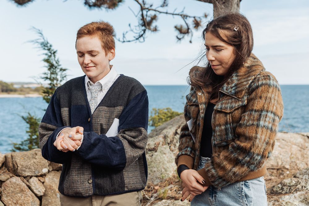 Two young women in plaid jackets stand on rocky coastal overlook with ocean and trees in background.