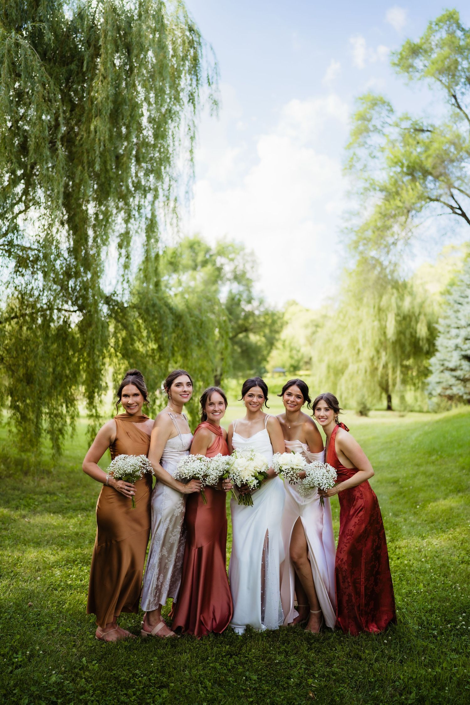 Bride and five bridesmaids in earth-tone satin gowns holding white bouquets outdoors near weeping willow trees.
