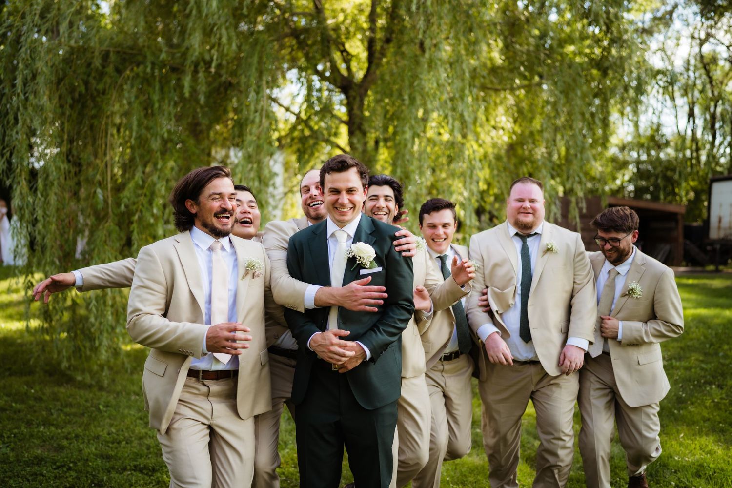 Groom in dark suit surrounded by groomsmen in tan suits celebrating outdoors under willow trees at a wedding.