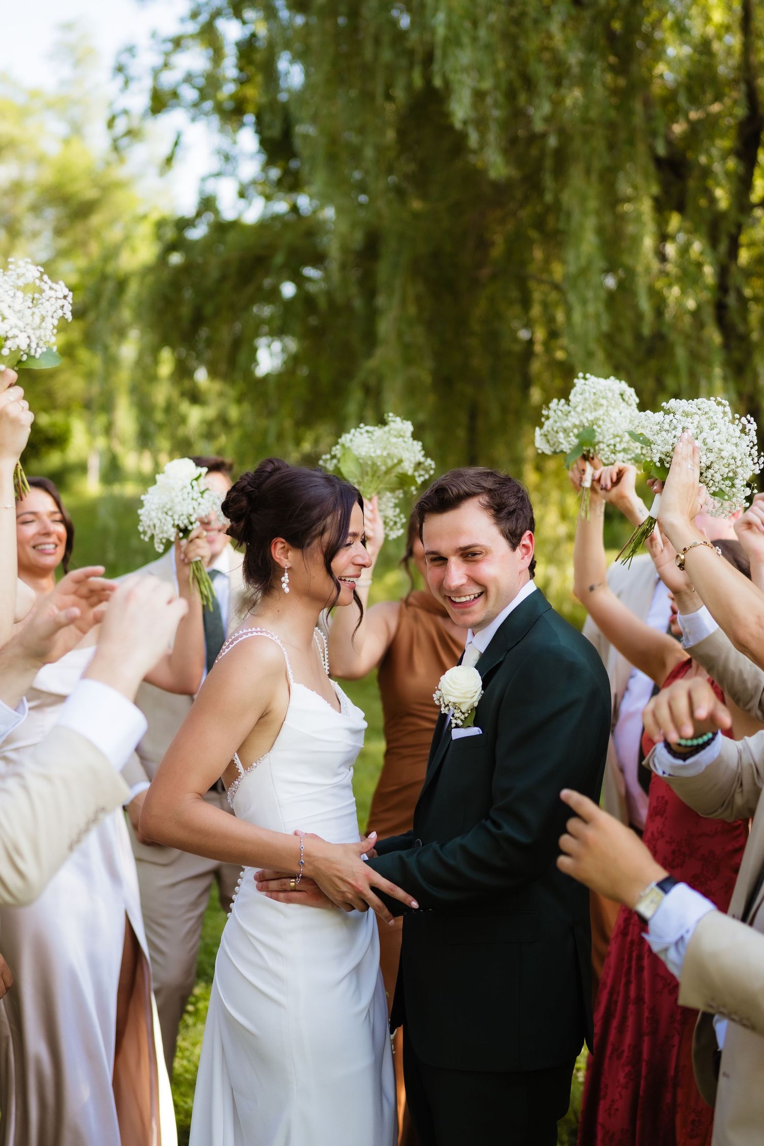 Happy bride and groom walk through cheering wedding guests holding white floral bouquets outdoors.