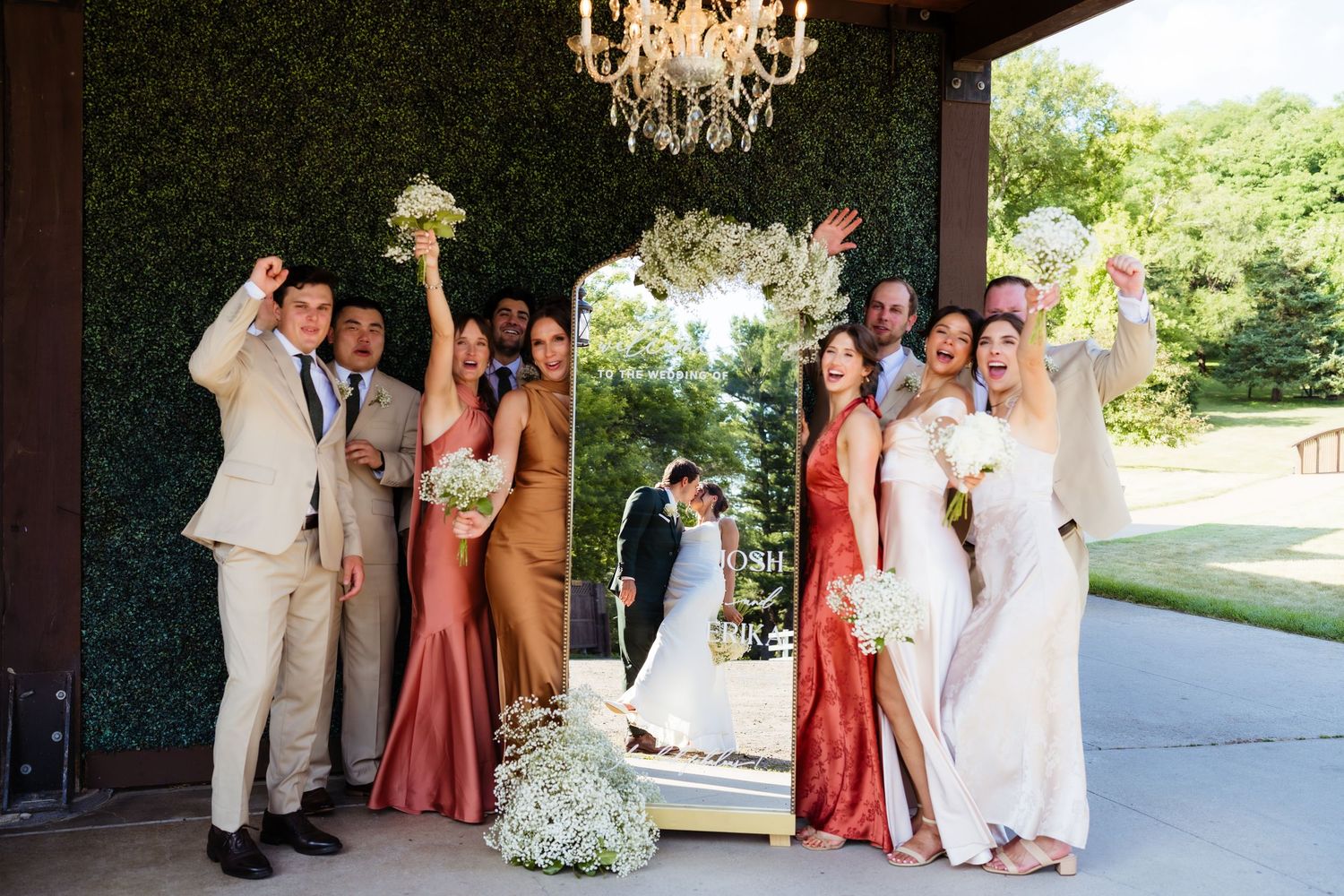 Wedding party celebrating beside a mirror as bride and groom kiss, with bridesmaids in gold and rust gowns outdoors.