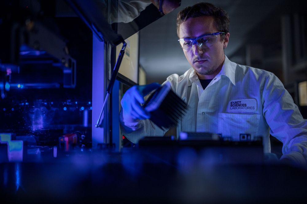 Scientist in white lab coat and blue gloves examines samples under blue laboratory lighting in research facility.