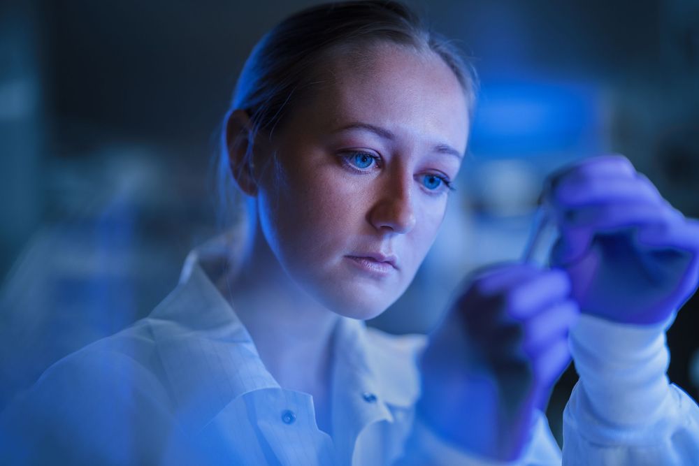 Female scientist in lab coat examining test tube under blue laboratory lighting in research facility.