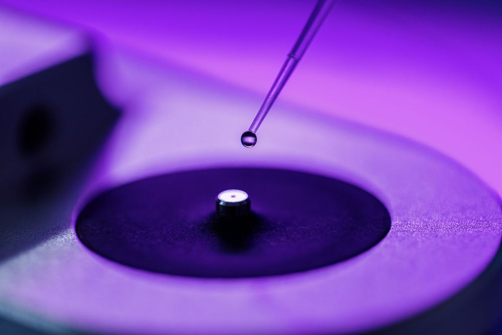 Close-up of a pipette dropping liquid onto a small lens component on a precision optical device under purple light.