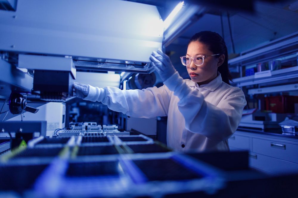 Female scientist in lab coat and safety goggles working with samples in a high-tech laboratory under blue lighting.