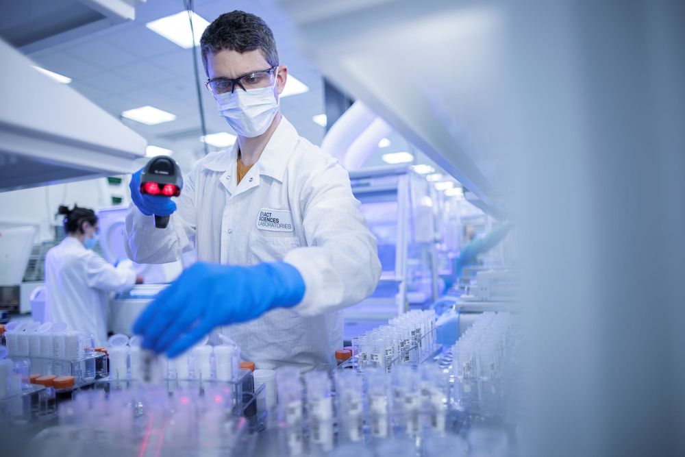 Lab technician in white coat and blue gloves handling samples in a modern pharmaceutical laboratory.