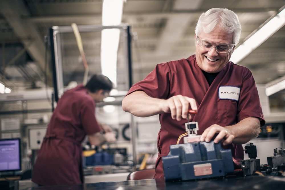 Smiling senior male technician in red uniform working with precision tools on industrial equipment in a manufacturing facility.