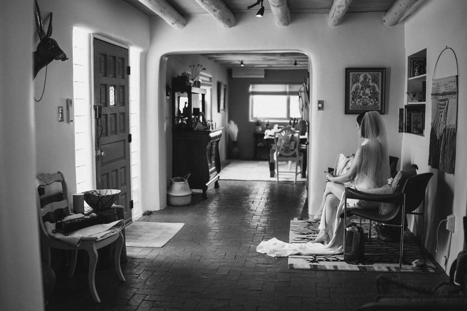 Black and white photo of a woman sitting in a rustic Southwestern-style hallway with adobe walls and tile floors.