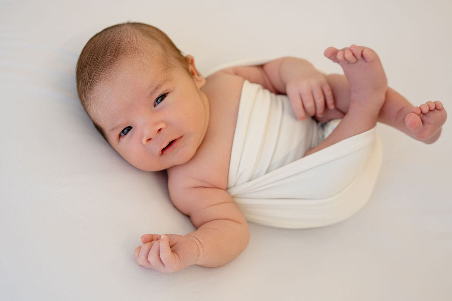 Newborn baby wrapped in white swaddle cloth lying on white background, looking up with bright eyes.