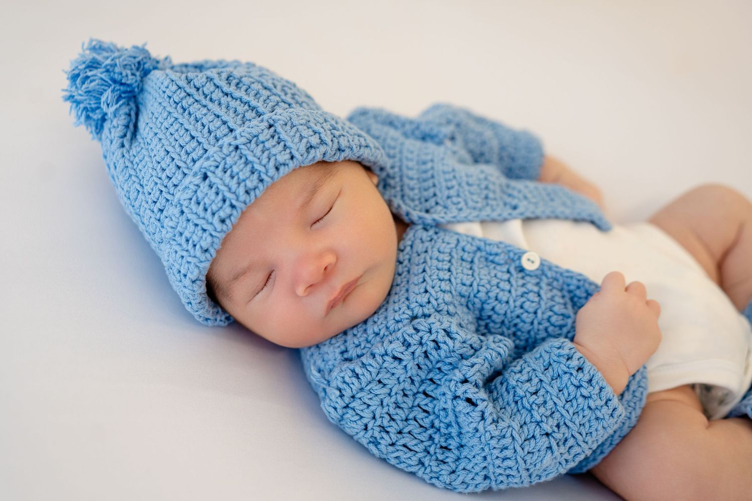 Sleeping newborn baby wearing a blue crochet knit hat and matching sweater outfit on a white background.