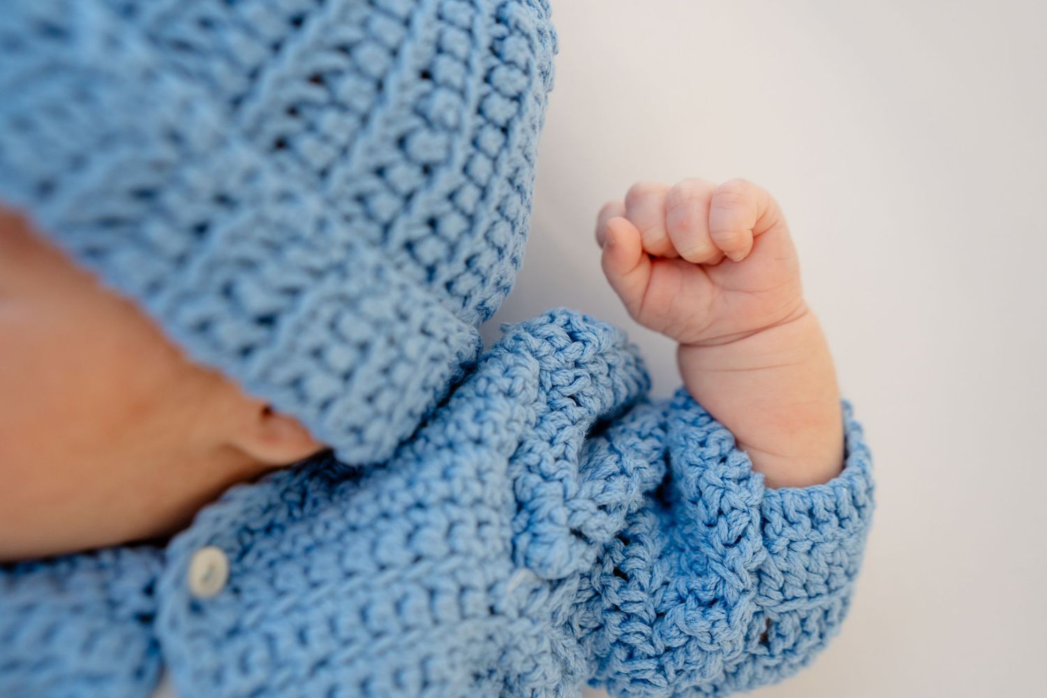 Close-up of a newborn baby's tiny fist in a blue crochet knit hooded sweater.