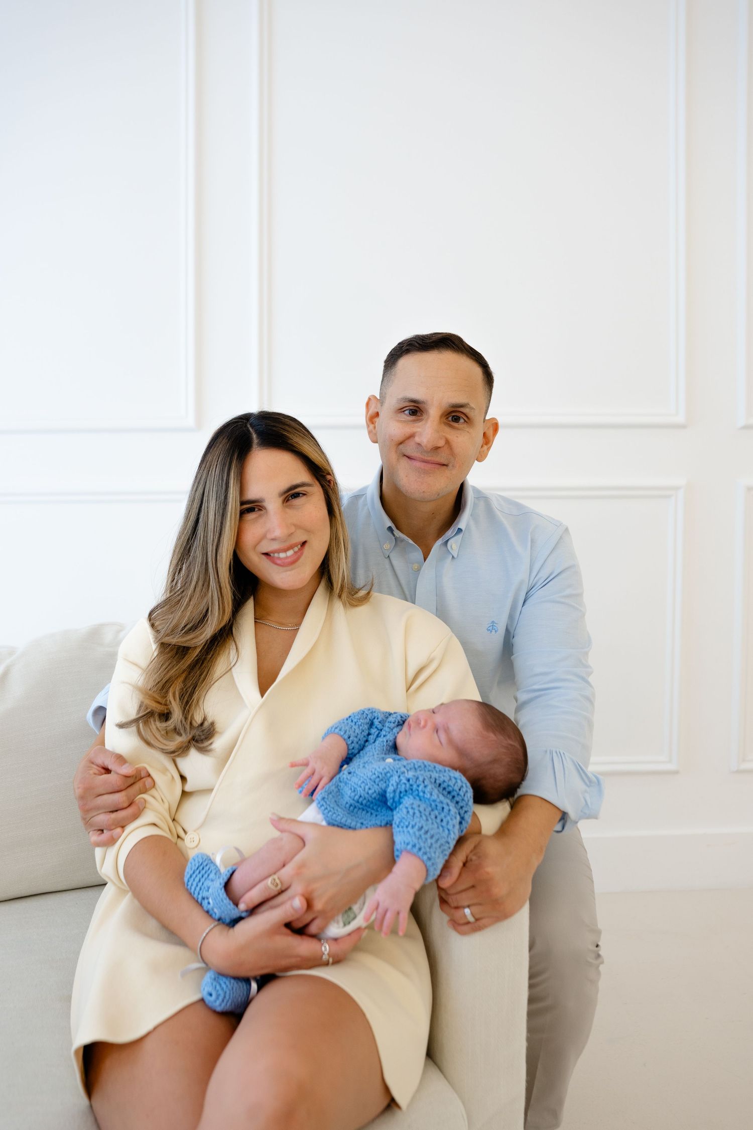 Happy couple sitting on a sofa holding their newborn baby dressed in a blue knit outfit.