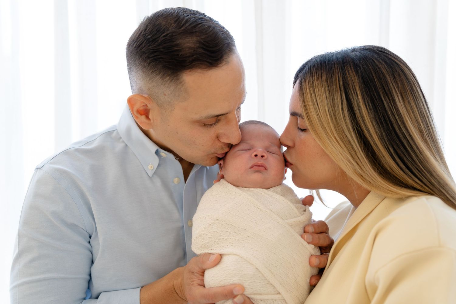 Parents kissing their newborn baby on the cheeks, wrapped in white swaddle, in a bright nursery room.