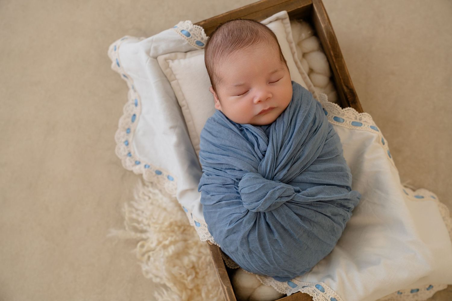 Newborn baby swaddled in blue wrap sleeping peacefully in a wooden box with white lace blanket.