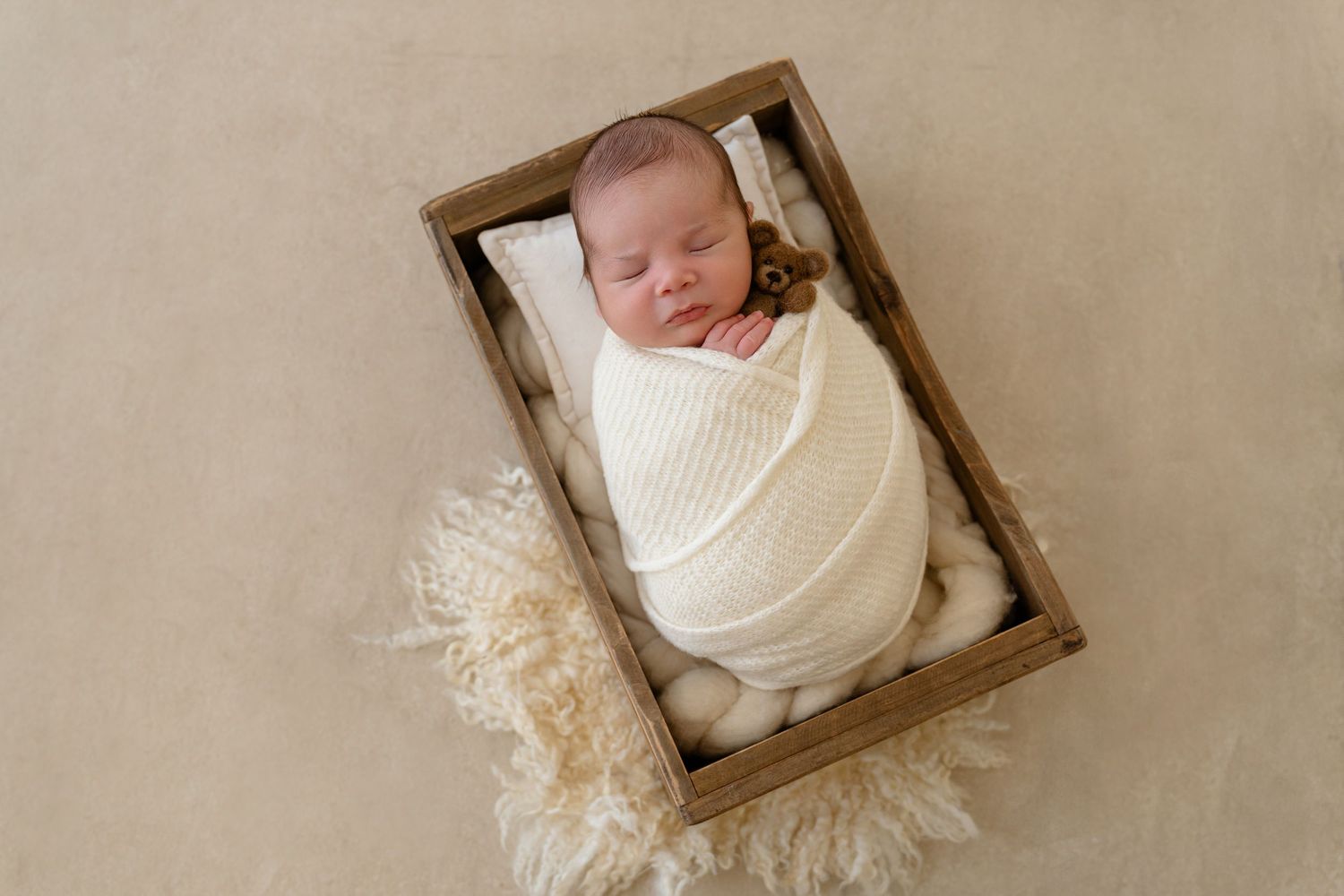 Newborn baby swaddled in cream wrap sleeping peacefully in rustic wooden crate with fluffy fur blanket.