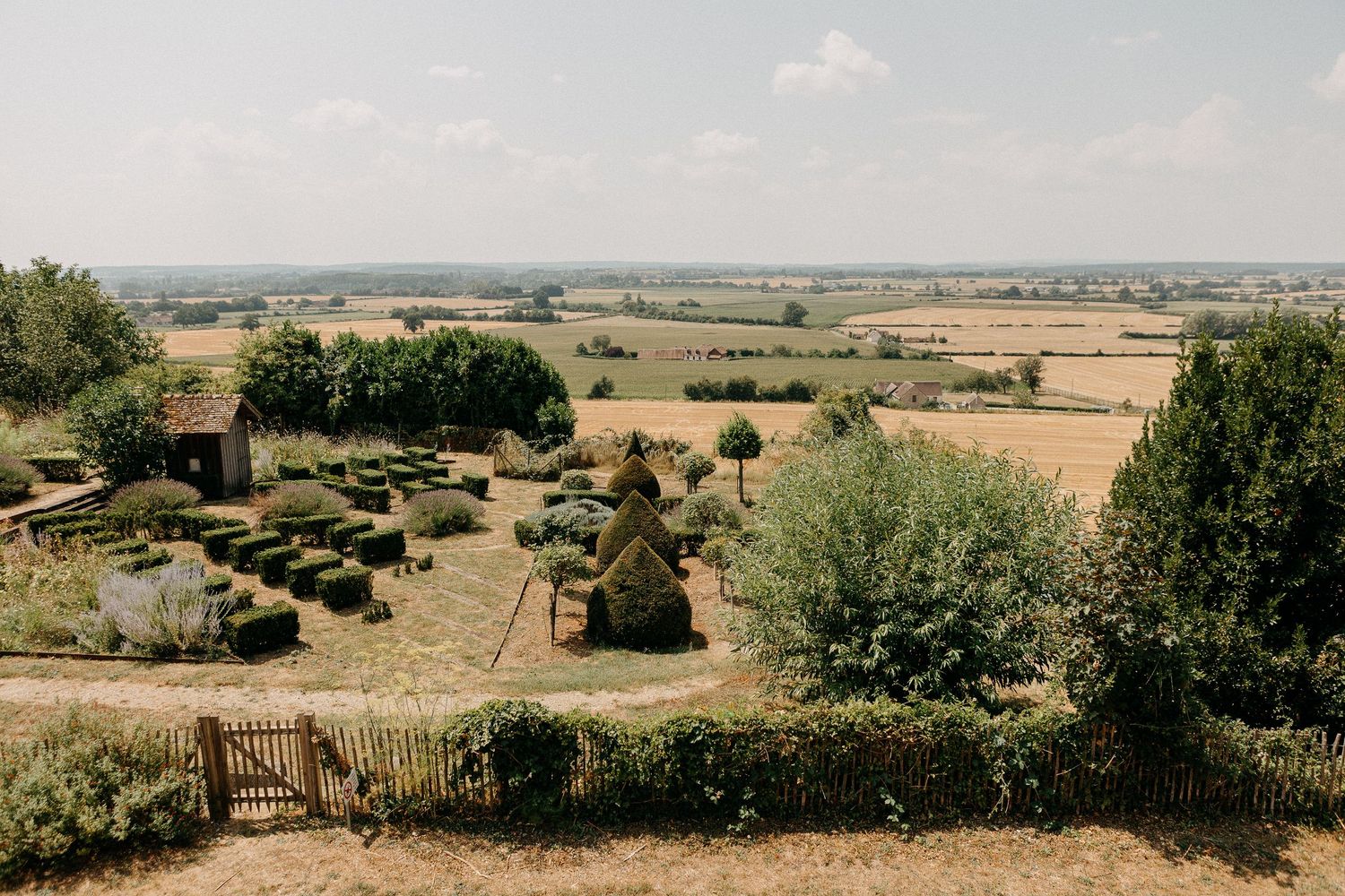 Aerial view of a formal English country garden with topiary hedges, overlooking vast golden fields under a clear blue sky.