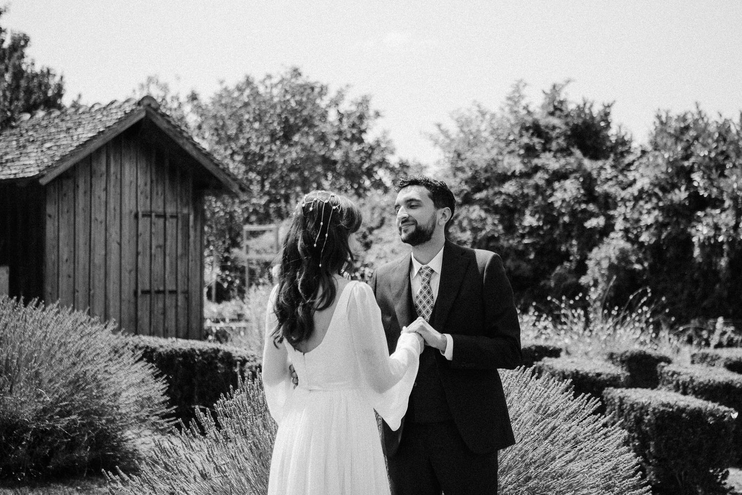 Black and white photo of a bride and groom sharing a joyful moment in a garden with lavender and a wooden shed.