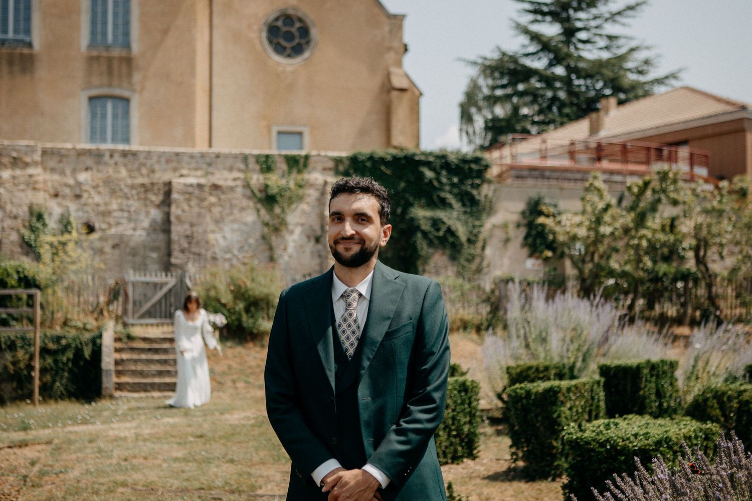 Groom in green suit smiling outdoors at wedding venue with bride in white dress visible in background.