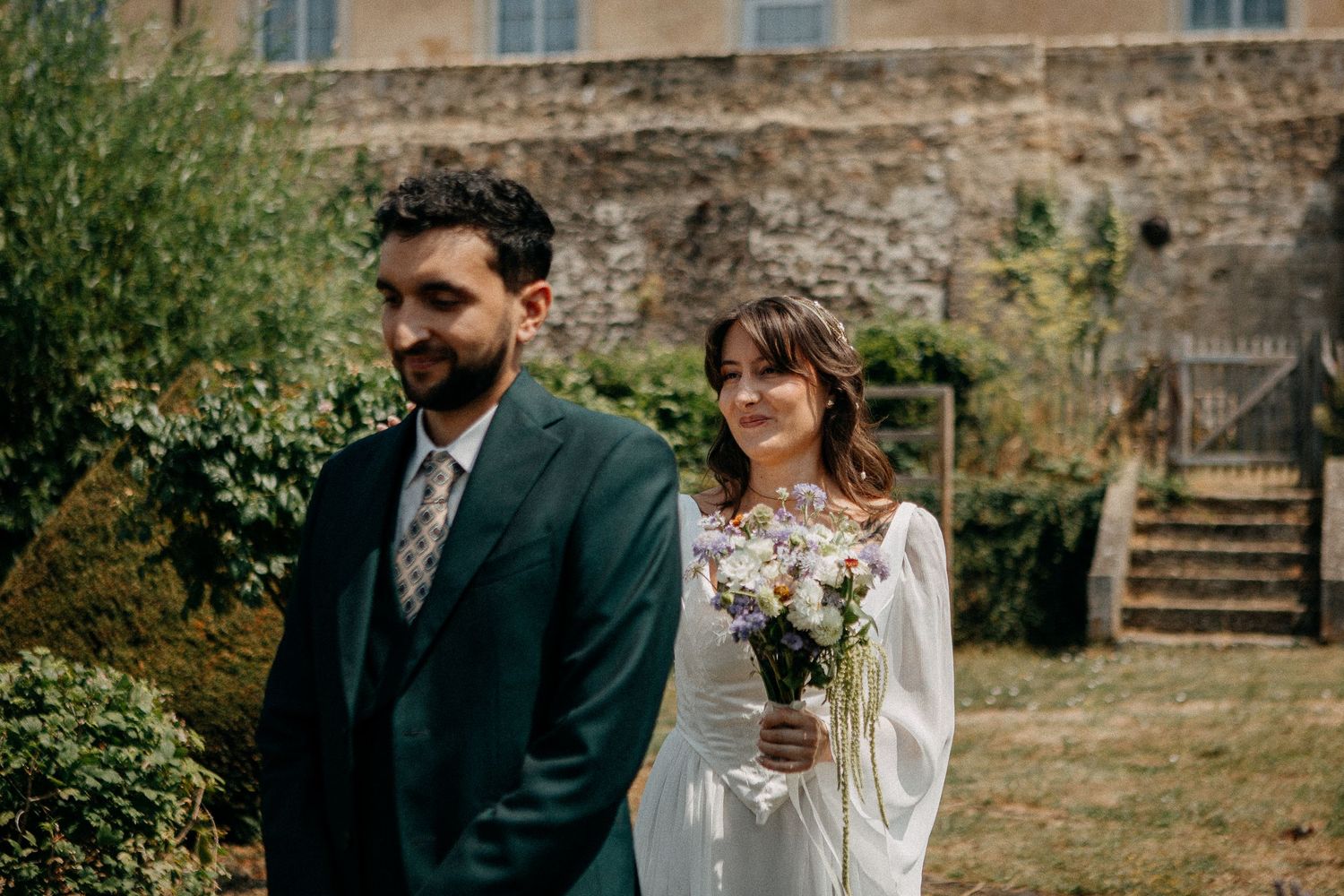 Bride holding wildflower bouquet stands behind groom in green suit at outdoor wedding venue with stone wall backdrop.