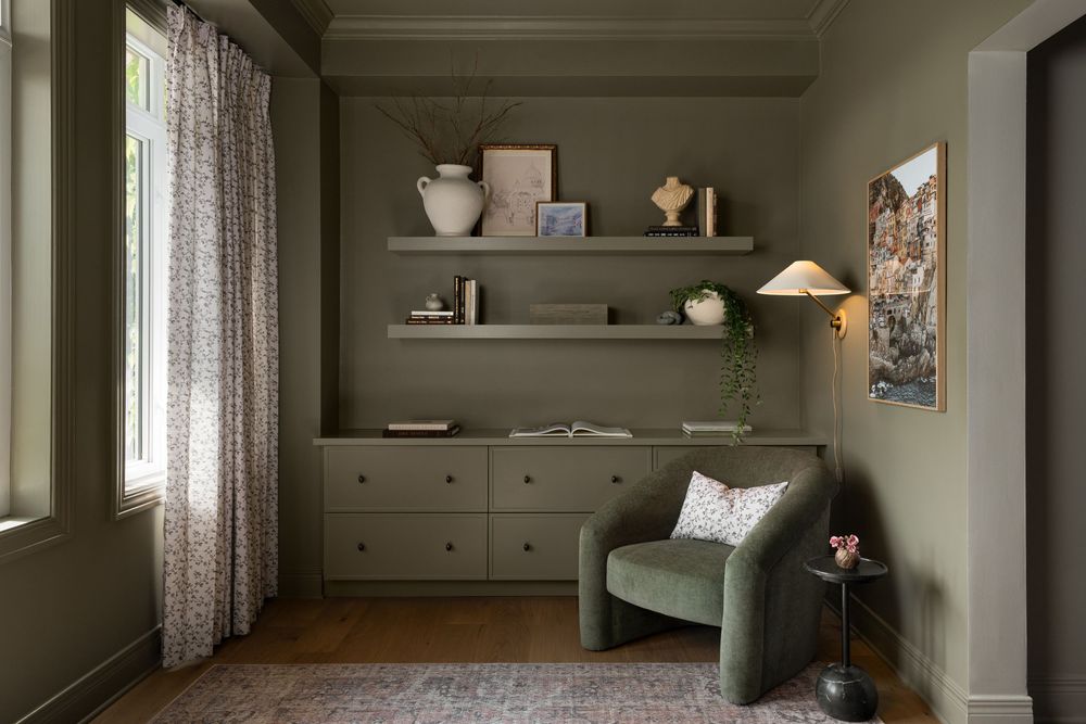Cozy olive green reading nook with built-in drawers, floating shelves, velvet armchair, and floral curtains in a stylish home.