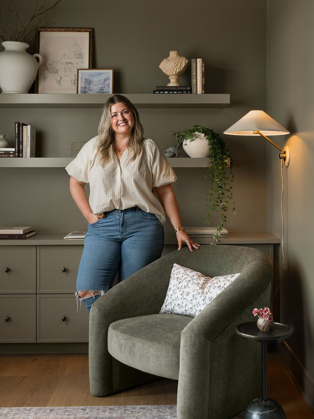 Woman smiling in a stylish olive green home office with floating shelves, an armchair, and a wall-mounted lamp.