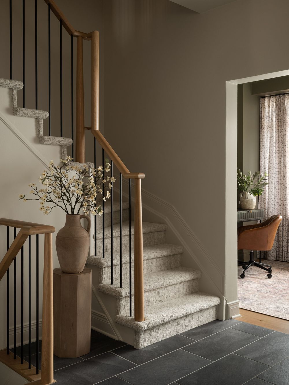 Elegant staircase with wood and metal railing, textured carpet steps, slate tile floor, and a neutral vase with white blooms.