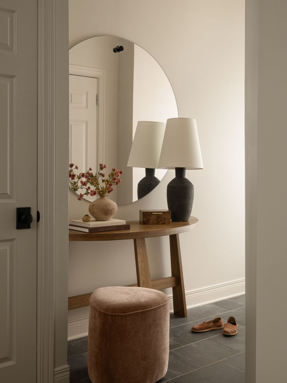 Minimalist entryway with arched mirror, wooden console table, black lamp, floral vase, and velvet ottoman on dark tile floor.