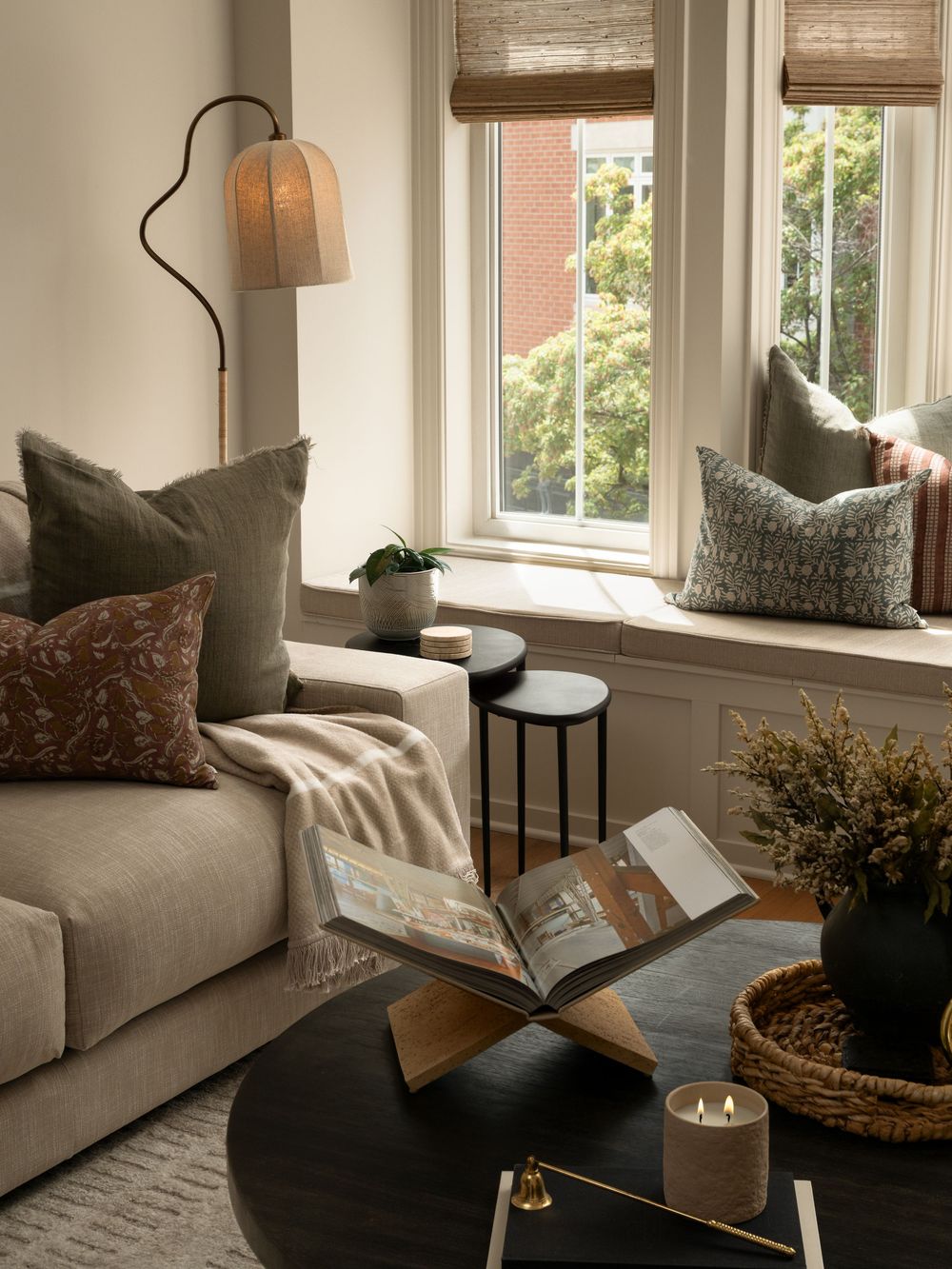 Cozy living room corner with beige sofa, earth-tone pillows, floor lamp, black coffee table, and window seat with green cushions.