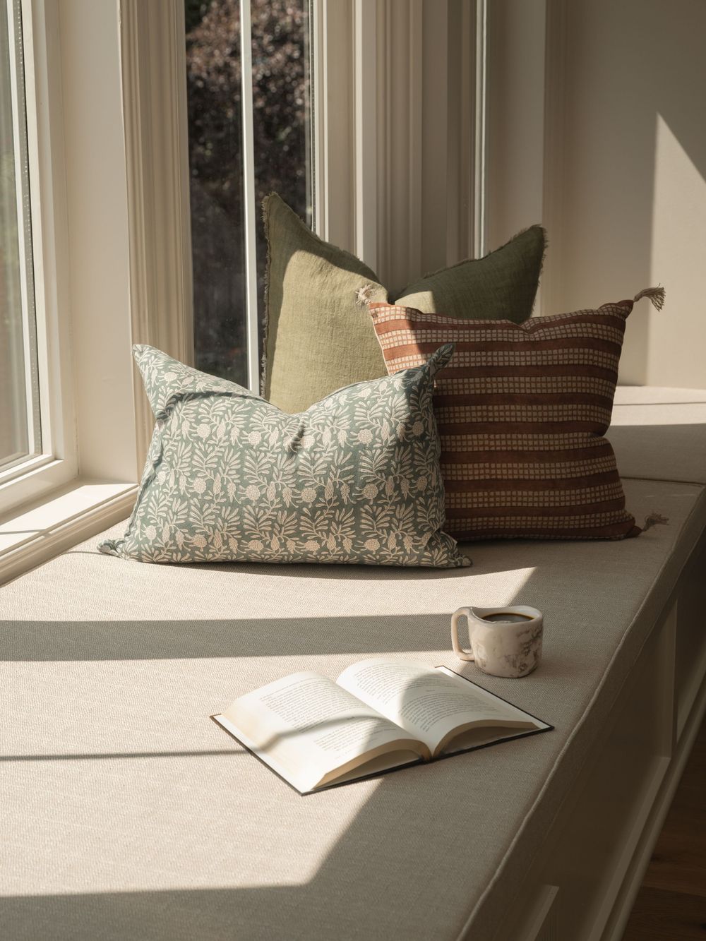 Cozy window seat with floral and striped throw pillows, an open book, and a mug in warm natural light.