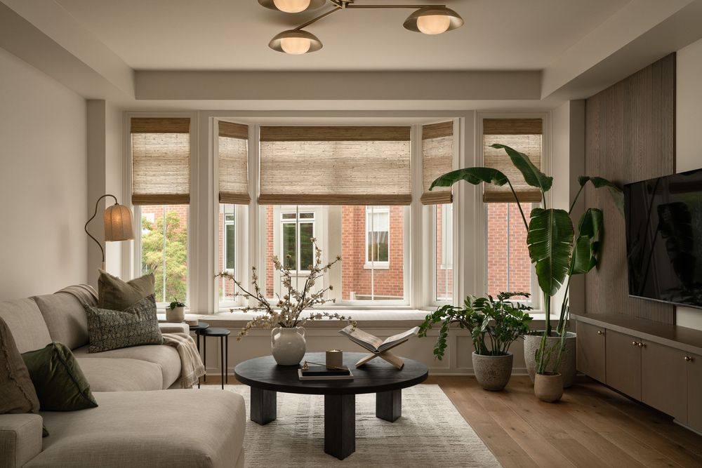 Modern living room with neutral tones, woven Roman shades, black coffee table, lush plants, and hardwood floors.