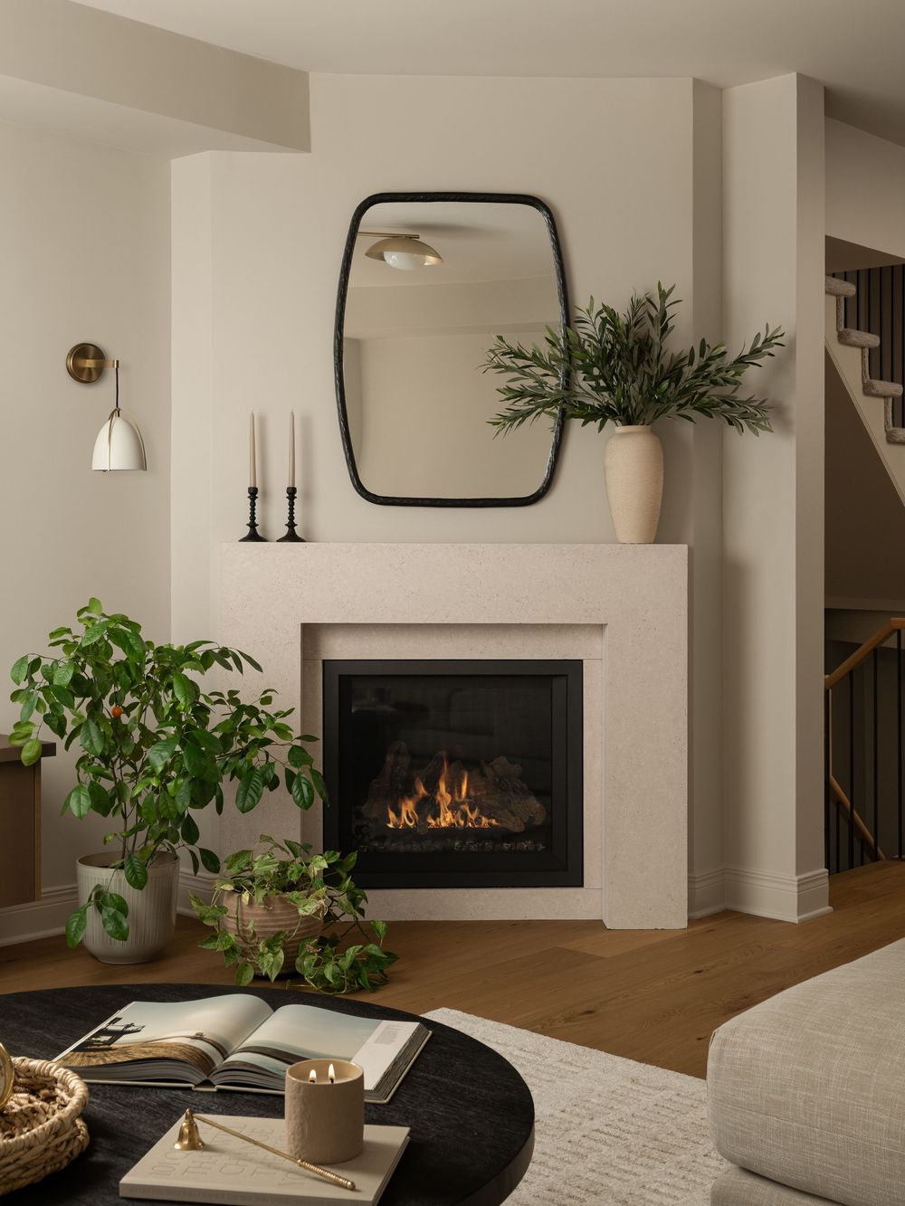 Cozy living room with lit fireplace, black mirror, candles, greenery, and warm wood floors.